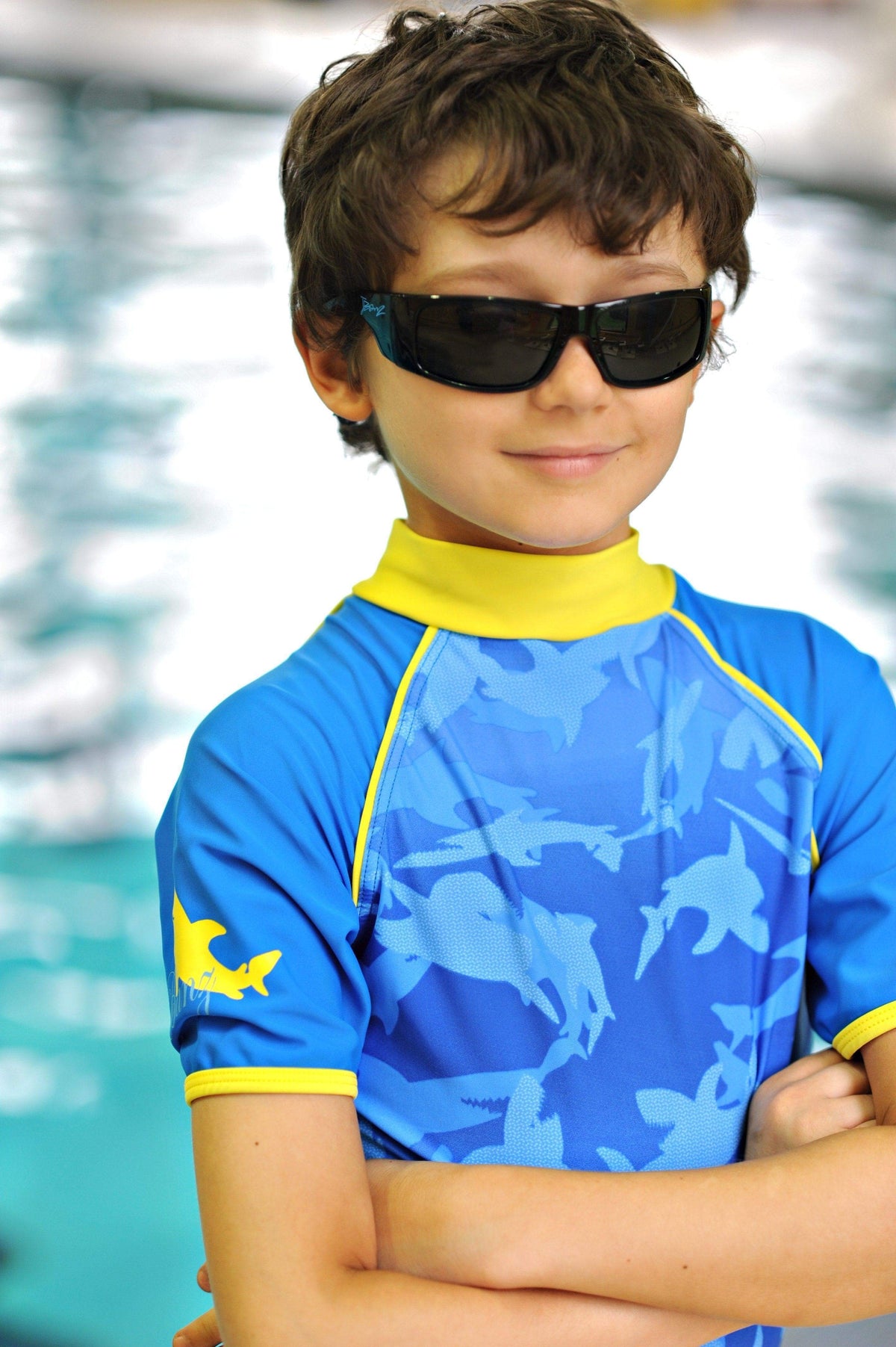 a school age boy crosses his arms and smiles at a pool wearing banz sunglasses and short sleeve rash top